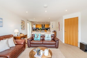 A living room with a brown leather sofa, a coffee table with a magazine, and a kitchen in the background. - 2 Bed in Kingswear (oc-bc129) (Waterhead)