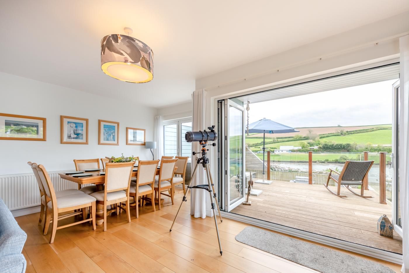 A dining area with a table and chairs, a camera on a tripod, and a view of a river and fields through large glass doors.
