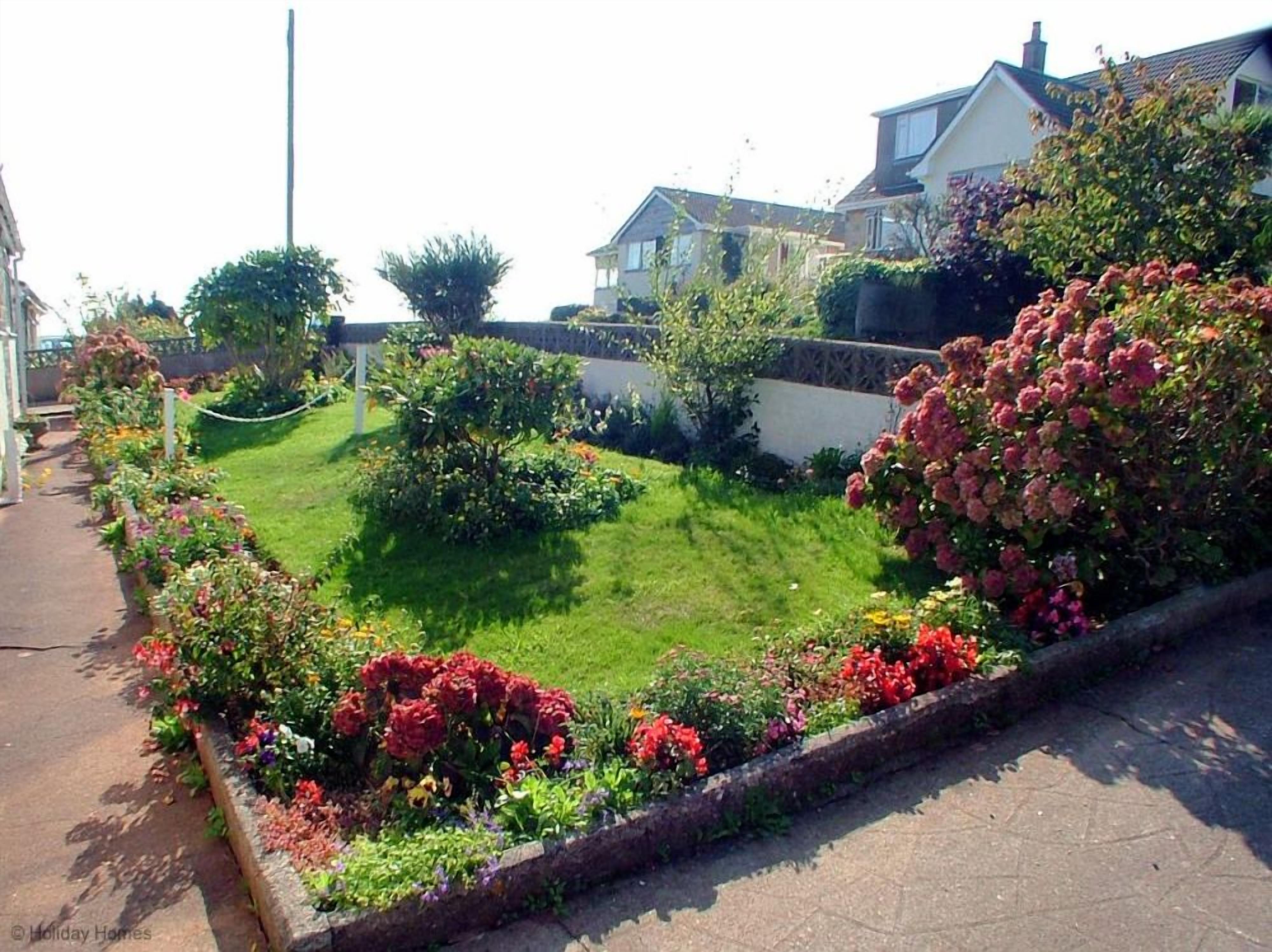 Um jardim residencial com gramado bem cuidado, diversas plantas floridas e um muro de pedra.