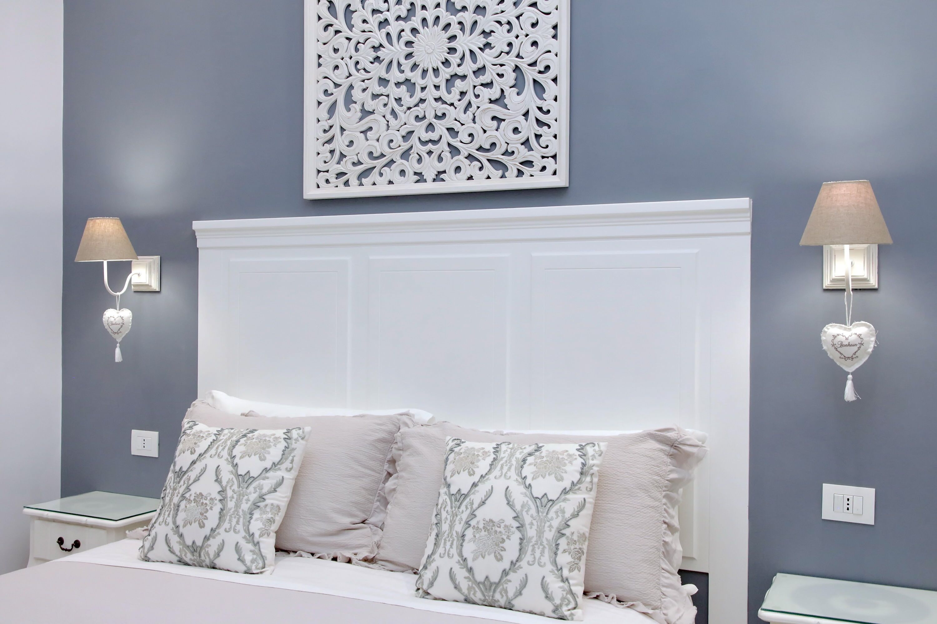 A bedroom with a white headboard, two bedside tables, a bed with patterned pillows, and wall-mounted lamps.
