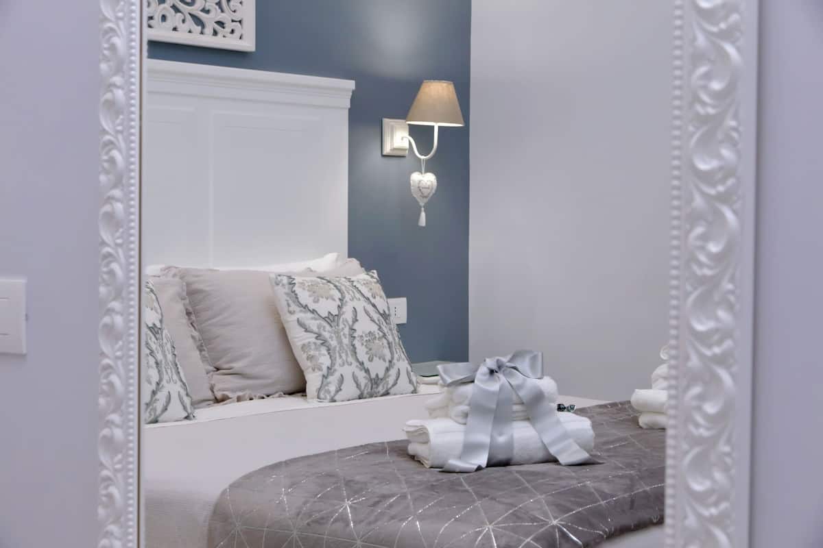 Un dormitorio con cama, almohadas, una lámpara de pared y un espejo decorativo.