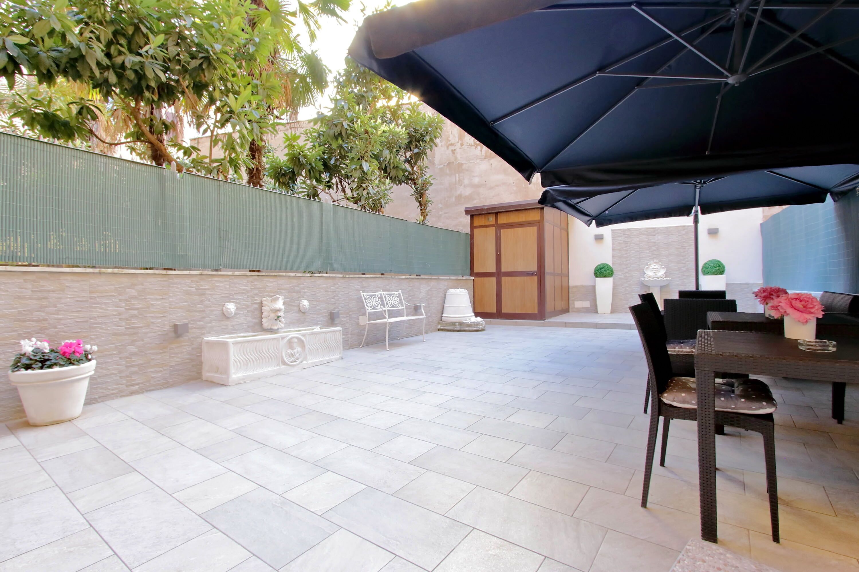 An outdoor patio area with a table and chairs, a large umbrella, potted plants, and a decorative bench.