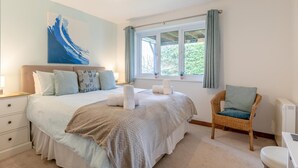 A bedroom with a large bed, a chair, a window with curtains, and a painting on the wall. - 4 Bed in Croyde (oc-sundow) (Croyde)