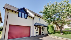 A two-story house with a red garage door, a small porch with a door, and a tree in the front yard. - 4 Bed in Croyde (oc-sundow) (Croyde)