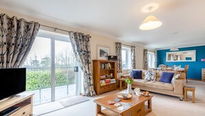 A modern living room with a sofa, coffee table, bookshelf, and a TV. - 4 Bed in Croyde (oc-sundow) (Croyde)