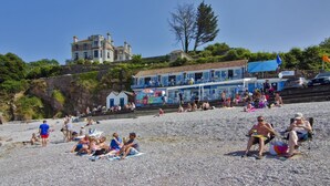 En strand med folk som solar och badar, en byggnad med en blå fasad och ett stort hus i bakgrunden.