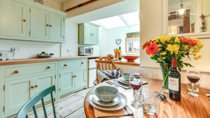 A dining area with a wooden table set for two, a vase of flowers, and a bottle of wine. - 2 Bed in Shaldon (oc-starco) (Shaldon)