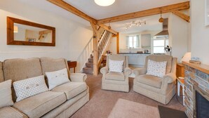 A living room with a sofa, two armchairs, a fireplace, a kitchen in the background, and a wooden staircase. - 2 Bed in Shaldon (oc-stchco) (Shaldon)