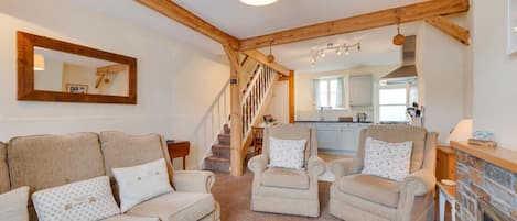 A living room with a sofa, two armchairs, a fireplace, a kitchen in the background, and a wooden staircase.
