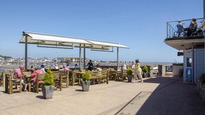 A seaside terrace with wooden benches and tables, a covered seating area, and a view of the sea and town in the background. - 2 Bed in Shaldon (oc-stchco) (Shaldon)