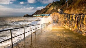 Une promenade en bord de mer avec une rambarde, une falaise rocheuse et un bâtiment en arrière-plan.
