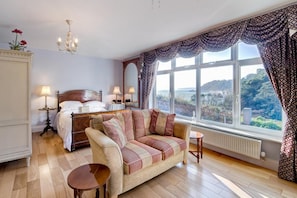 A bedroom with a large bed, a sofa, a chandelier, and a view of the outdoors.