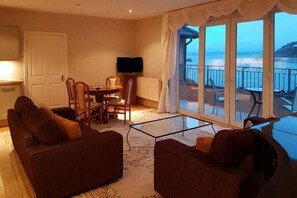 A living room with a sofa set, a dining table with chairs, a television, and a large sliding glass door leading to a balcony with a view of the sea.