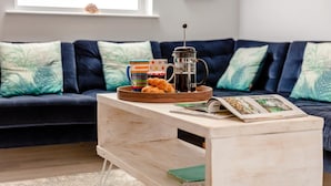 A cozy living room with a dark blue sofa, a wooden coffee table, and a window with a view. - 4 Bed in Shaldon (oc-driftc) (Shaldon)