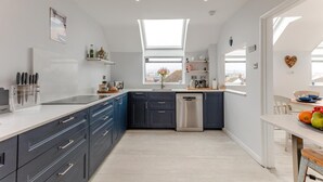 A modern kitchen with dark cabinets, a stainless steel dishwasher, and a large window providing natural light. - 4 Bed in Shaldon (oc-driftc) (Shaldon)