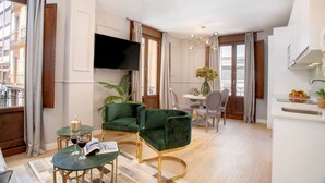 A modern living room with a green sofa, a coffee table, and a TV mounted on the wall. There is a dining area with a table and chairs, and a kitchen area with a sink and countertop. - Genteel Home Mesones I (Granada)