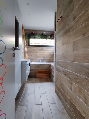 A narrow bathroom with a bathtub, wooden floor, and a window with plants.