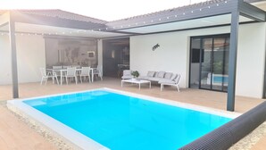 Ett modernt utomhusområde med en pool med kristallklart blått vatten, en täckt uteplats med ett matbord och stolar samt en soffa.