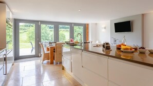 A modern kitchen with a large island, a dining area with wicker chairs, and a view of greenery through large sliding doors. - 4 Bed in Drewsteignton (oc-bc2408) (Drewsteignton)