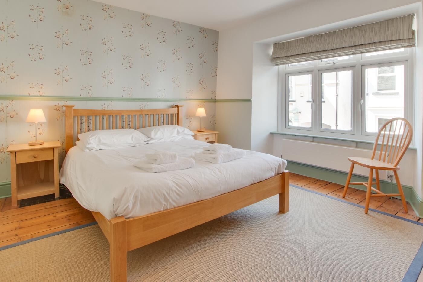 Une chambre à coucher avec un lit en bois, deux tables de chevet avec des lampes, une chaise et une fenêtre avec des stores.