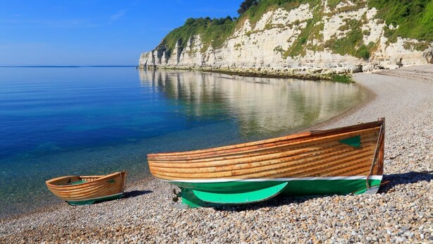 Une plage de galets avec deux bateaux, une falaise et un ciel bleu dégagé.