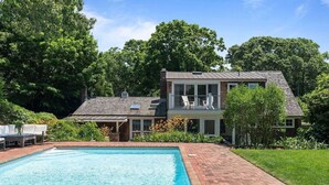 A two-story house with a swimming pool, surrounded by lush greenery. - Classic Hamptons home in Springs.  Pool, lovely back yard and steps to the beach (East Hampton)