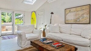 A modern living room with white sofas, a coffee table with books, a surfboard, and a map on the wall. - Classic Hamptons home in Springs.  Pool, lovely back yard and steps to the beach (East Hampton)