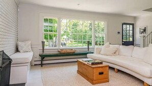 A living room with a white sofa, a wooden coffee table with books, a bench with cushions, and large windows with a view of greenery. - Classic Hamptons home in Springs.  Pool, lovely back yard and steps to the beach (East Hampton)