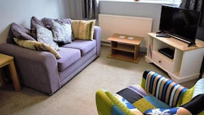 Una sala de estar con sofá, televisión, una pequeña mesa de madera y una silla colorida.