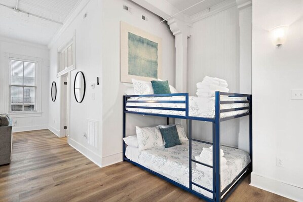 A bunk bed with a blue frame and white bedding, a painting on the wall, and a window with blinds.