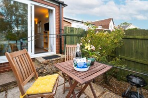An outdoor patio with a wooden table, chairs, a bottle, and a vase of flowers.