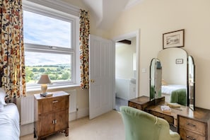 A bedroom with a large window, a wooden dresser, a green armchair, a bedside table with a lamp, and a mirror.