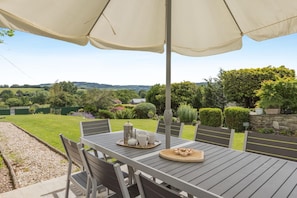An outdoor patio with a table set for tea, surrounded by chairs, and an umbrella providing shade.