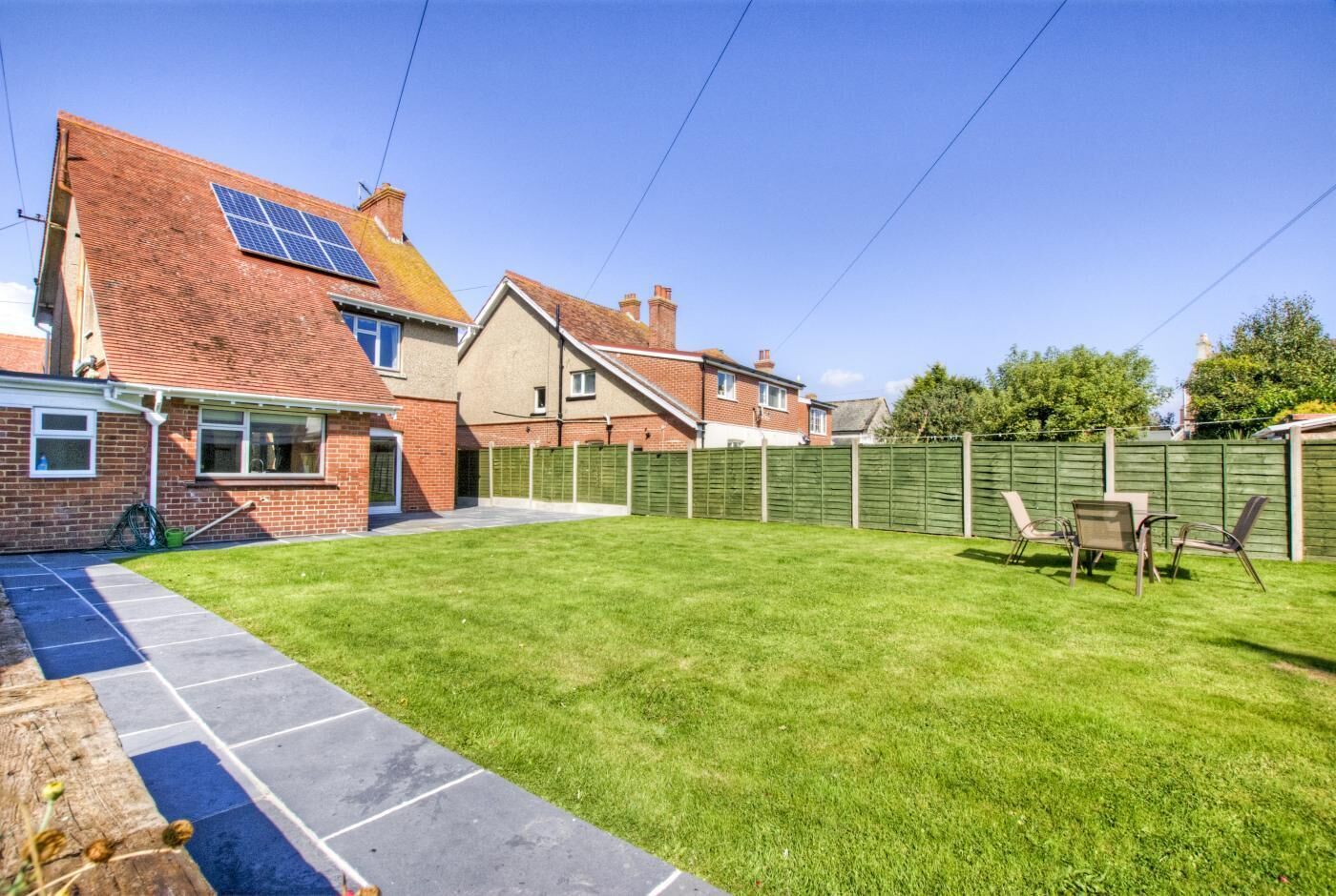 Ein Wohnhof mit gepflasterter Terrasse, grüner Rasenfläche, Holzzaun und einem Solarmodul am Haus.
