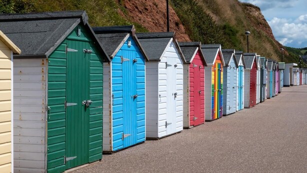 A row of colorful beach huts with sloped roofs, lined up along a paved walkway. - 3 Bed in Seaton (oc-mbdal) (Seaton)