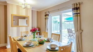 A dining area with a table set for four, chairs, a vase with flowers, and a view through a glass door to an outdoor patio. - 3 Bed in Seaton (oc-mbdal) (Seaton)