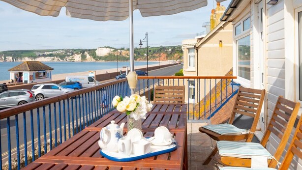 Un balcon avec une table préparée pour le thé, une chaise et une vue sur la mer et les bâtiments.