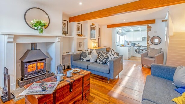 A cozy living room with a wood-burning stove, a sofa, a coffee table, and a kitchen in the background. - 2 Bed in Branscombe (oc-mbhave) (Branscombe)