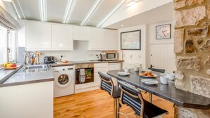 A modern kitchen with white cabinets, a stone accent wall, a built-in oven, and a dining area with bar stools. - 2 Bed in Branscombe (oc-mbhave) (Branscombe)
