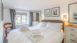 A neatly made bed with white linens and towels, a wooden chair, a bedside table with a lamp, and framed pictures on the wall. - 2 Bed in Branscombe (oc-mbhave) (Branscombe)