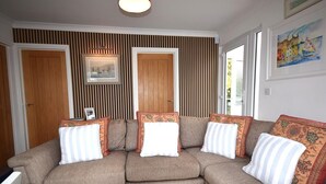 A living room with a sectional sofa, a framed picture on the wall, and a window with a view. - 2 Bed in Teignmouth (oc-bchyvi) (Teignmouth)
