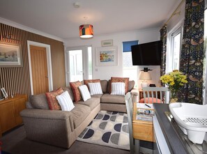 A modern living room with a sectional sofa, a wooden cabinet, a television, and a window with curtains. - 2 Bed in Teignmouth (oc-bchyvi) (Teignmouth)