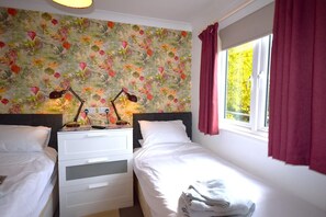A bedroom with two beds, a nightstand with lamps, a window with curtains, and a floral wallpaper. - 2 Bed in Teignmouth (oc-bchyvi) (Teignmouth)