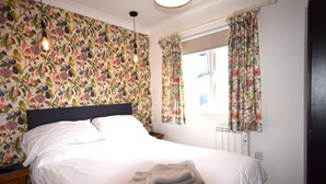 A neatly made bed with white linens and a folded towel, a nightstand with a lamp, and a window with floral curtains. - 2 Bed in Teignmouth (oc-bchyvi) (Teignmouth)