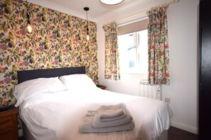 A neatly made bed with white linens and a folded towel, a nightstand with a lamp, and a window with floral curtains. - 2 Bed in Teignmouth (oc-bchyvi) (Teignmouth)