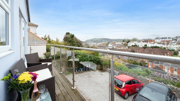 A balcony with wicker furniture, a glass railing, and a view of a residential area with cars and houses. - 2 Bed in Teignmouth (oc-bchyvi) (Teignmouth)