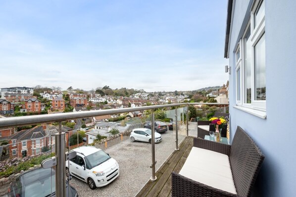 A balcony with a view of a residential area, a white car parked on the street, and a table with a vase of flowers.