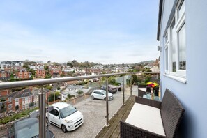 A balcony with a view of a residential area, a white car parked on the street, and a table with a vase of flowers.