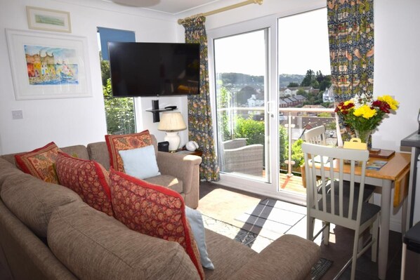 A living room with a sofa, a TV, a table with flowers, and a view of the outdoors. - 2 Bed in Teignmouth (oc-bchyvi) (Teignmouth)
