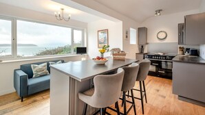 A modern kitchen with a large island, a dining area with bar stools, a sofa, and a view of the sea.